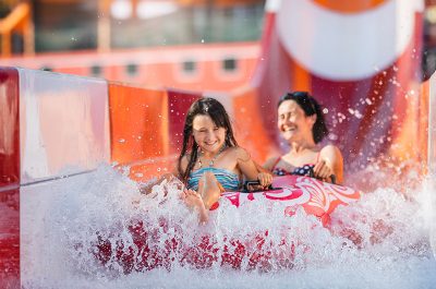 Madre e hija sonriendo mientras descienden juntas por un tobogán acuático con flotador, salpicando agua en Aquamijas, ideal para celebrar el Día de la Madre en Málaga.